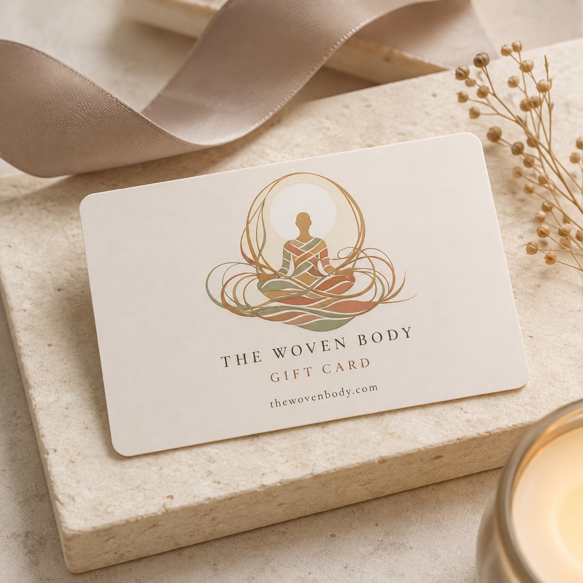 A cream Woven Body gift card on a stone tile, beside a satin ribbon, dried flowers and a lit candle